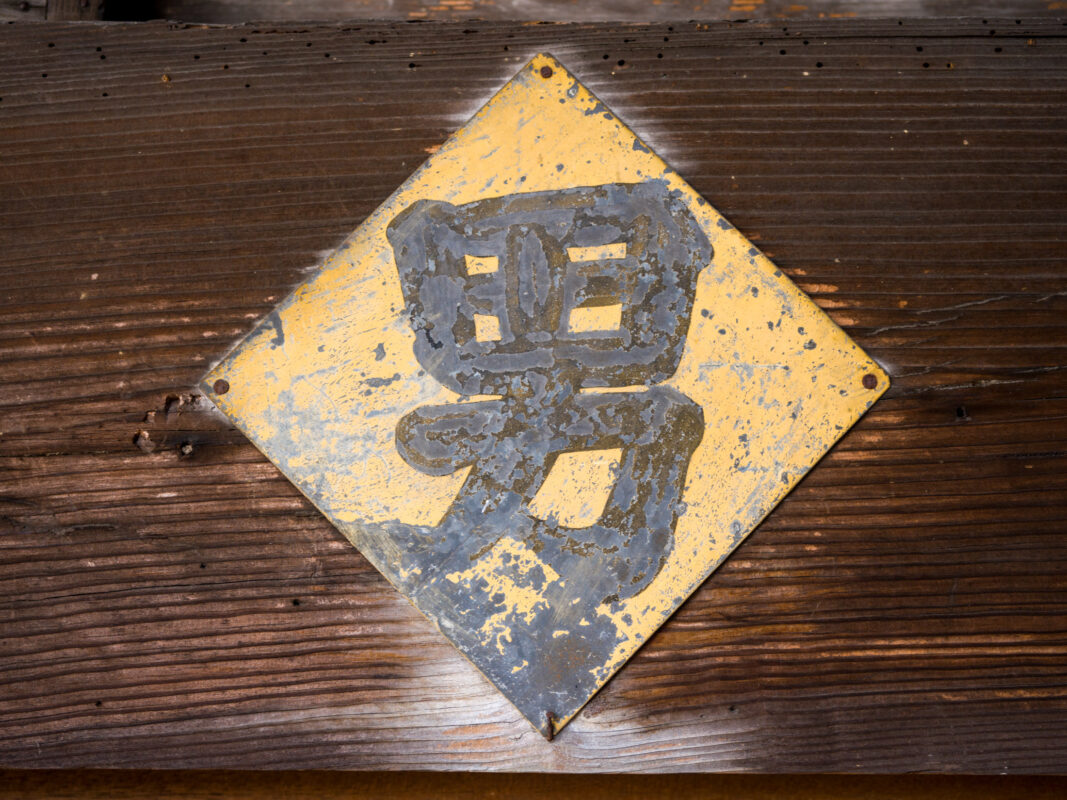 Weathered yellow Japanese kanji “Meguri” plaque on dark wooden boards, chipped vintage sign