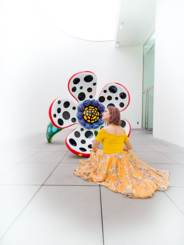 Visitor seated in Yayoi Kusama Museum gallery facing large polka-dot flower sculpture.
