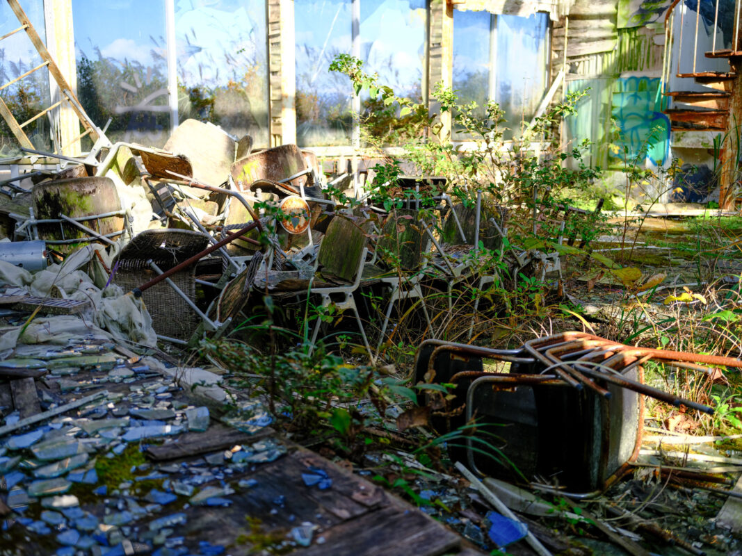 Sunlight through broken church windows over collapsed pews, rubble, and weeds reclaiming the interior.