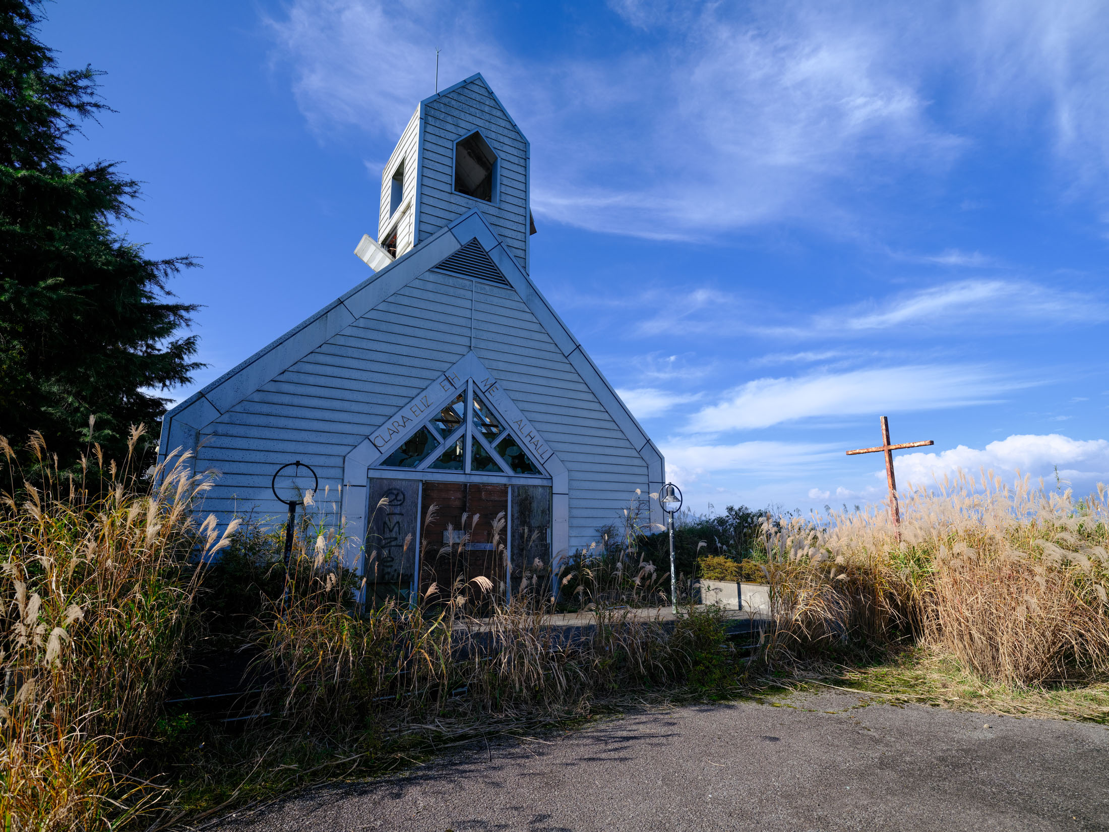 Forgotten rural church on overgrown path under blue sky, wooden cross nearby