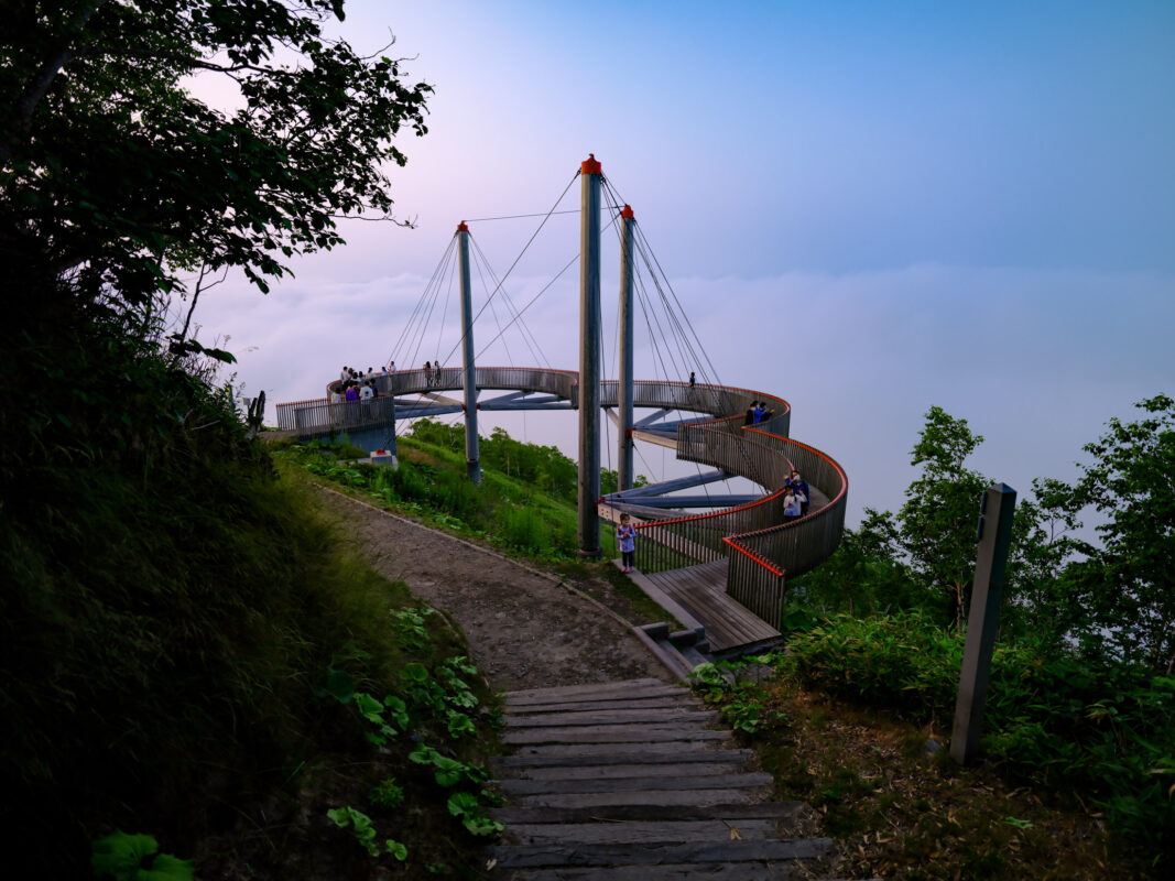 Curved mountain skywalk terrace above sea of clouds with visitors and cable supports.