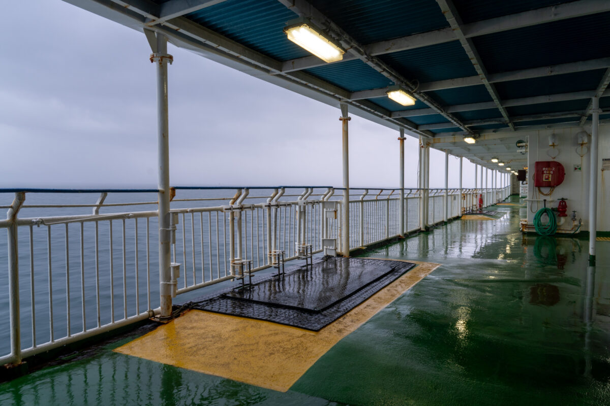 Rain-slick ferry deck walkway with railings overlooking calm ocean under overcast sky