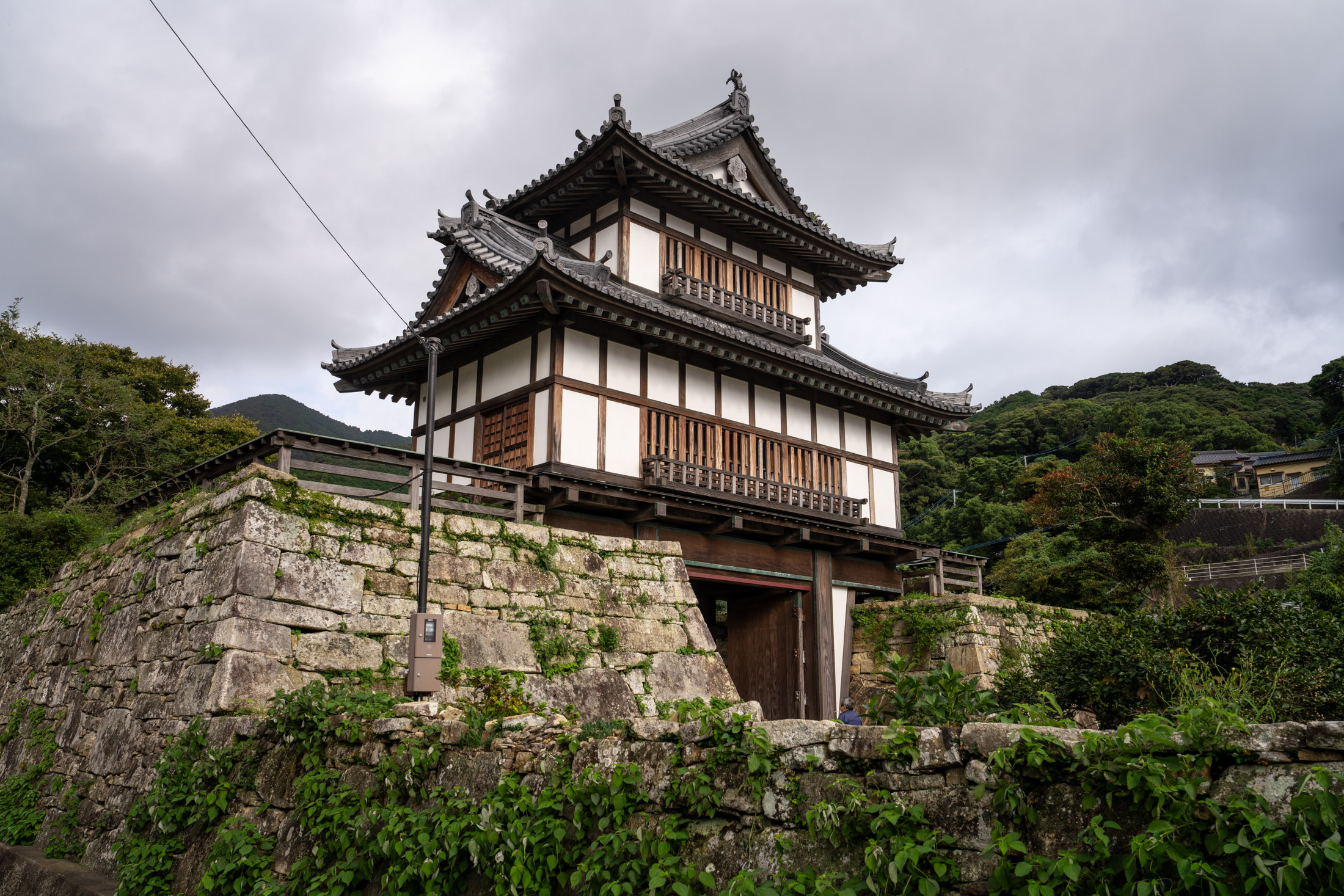 Historic Japanese gate tower on stone wall in Izuhara under cloudy sky