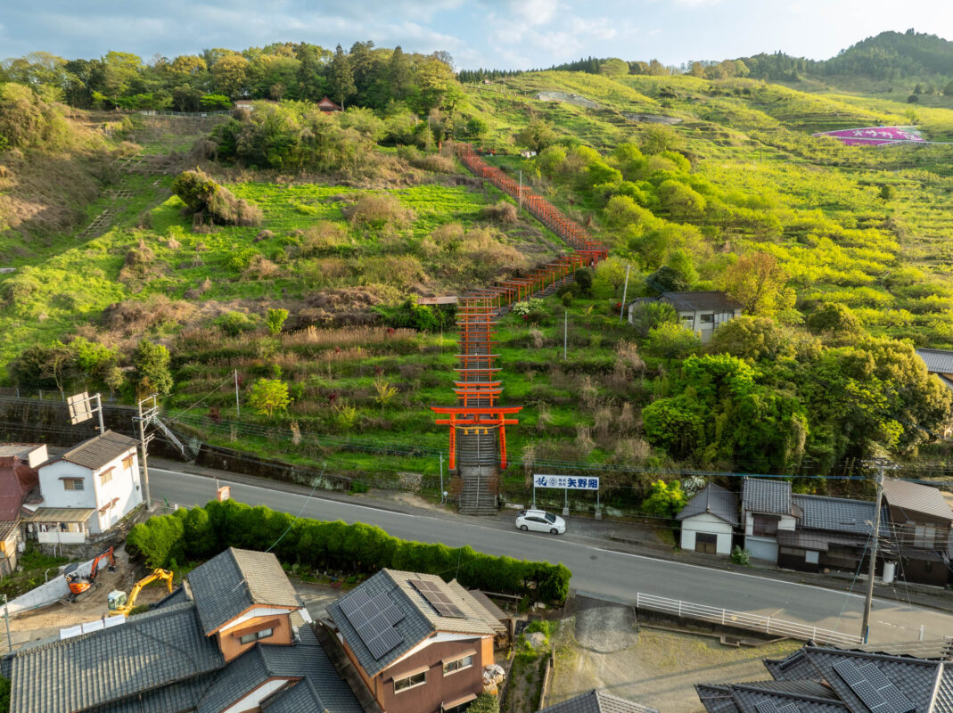 Vermilion torii gates climb Ukiha Inari Shrine hillside above rural Japan road and houses.