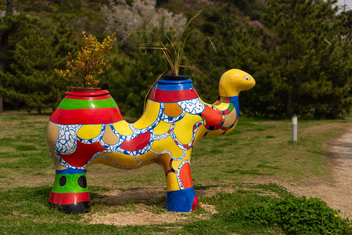 Colorful mosaic camel planter sculpture with grasses, displayed outdoors in Naoshima art park.