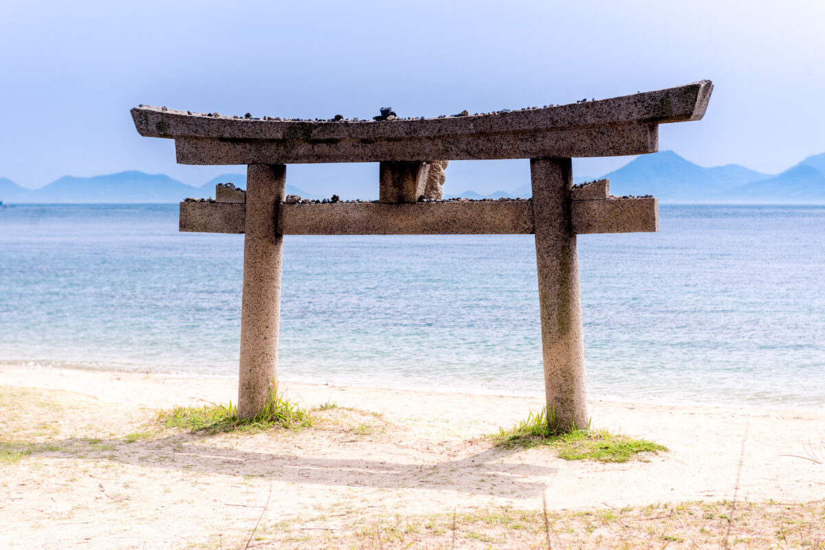 Weathered stone torii gate on Naoshima beach overlooking calm sea and distant mountains.