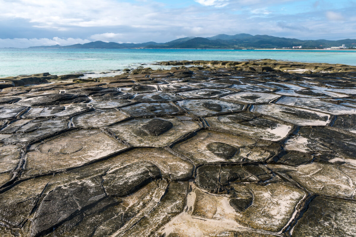 Kumejima rocky shoreline with polygonal stone slabs and clear turquoise sea under cloudy sky.