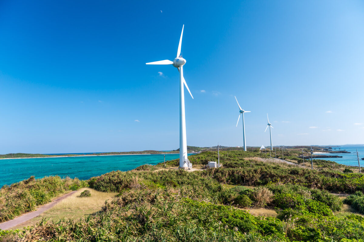 White wind turbines on Miyako Island coastline above turquoise ocean under clear blue sky.