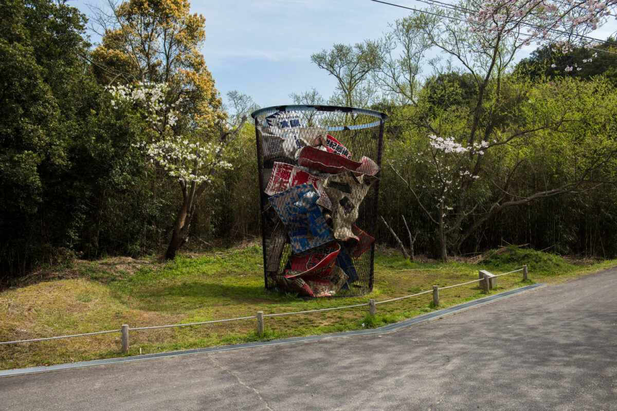 Cylindrical metal mesh sculpture filled with colorful debris along a path in Naoshima, Japan.