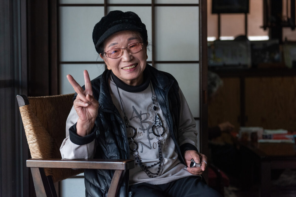 Smiling person in traditional Japanese room gives peace sign, wearing cap and glasses.
