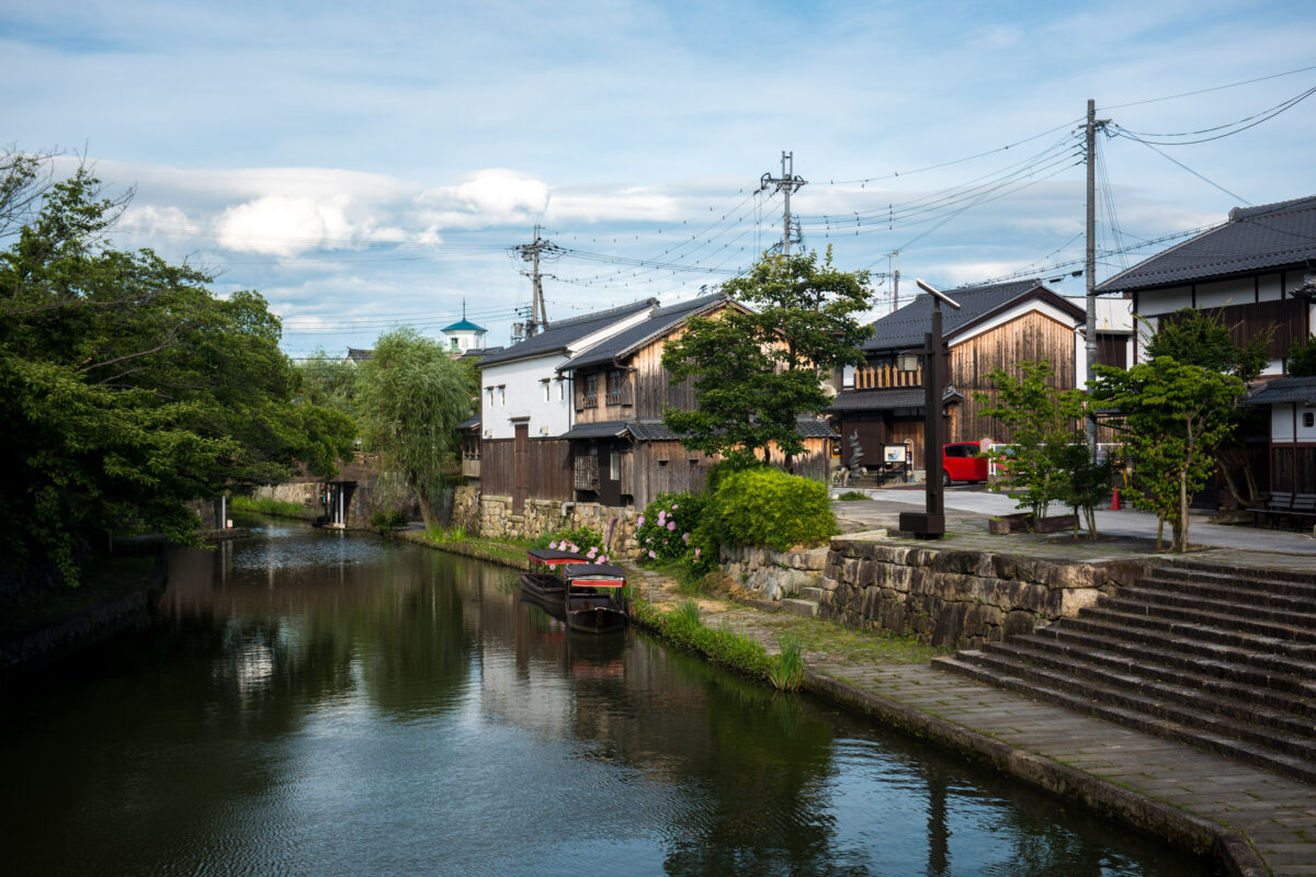 Omihachiman canal in Shiga with historic wooden townhouses, stone steps, trees, and boat.