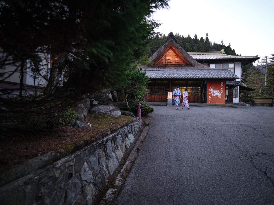 Glowing Shima Onsen ryokan at dusk in forested mountains, guests in yukata walking