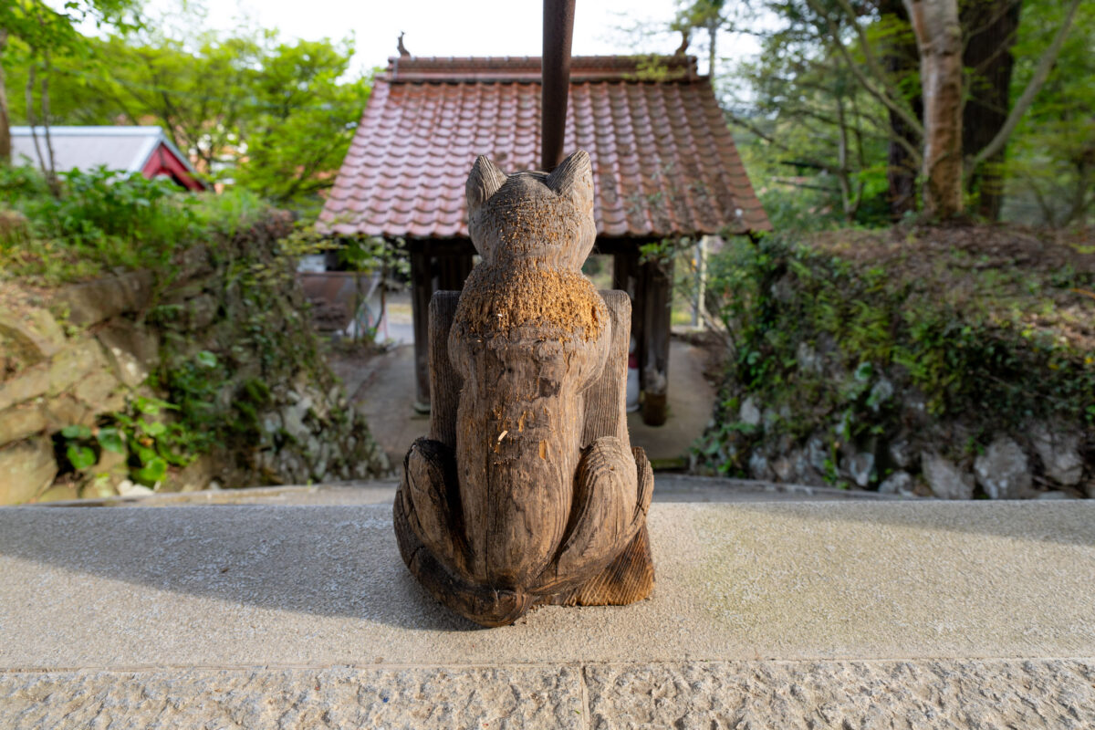 Weathered wooden fox statue overlooks Unrin-ji temple gate amid lush greenery