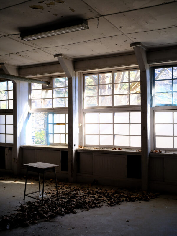 Abandoned classroom with dusty windows, fallen leaves, and lone desk, Silent Hill atmosphere.