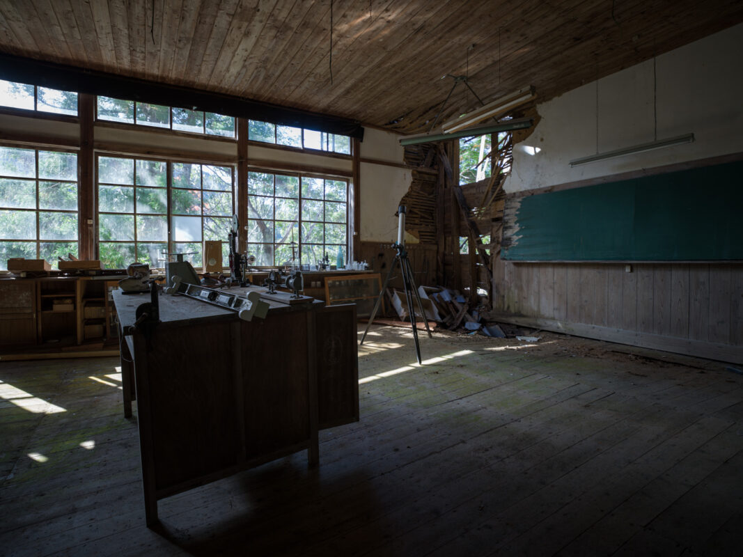 Sunlit abandoned classroom with broken wall, dusty desks, and forest views through windows.
