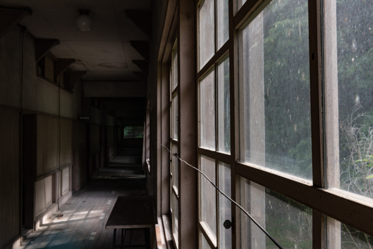 Abandoned school hallway with grimy windows, dusty floors, and eerie muted daylight.