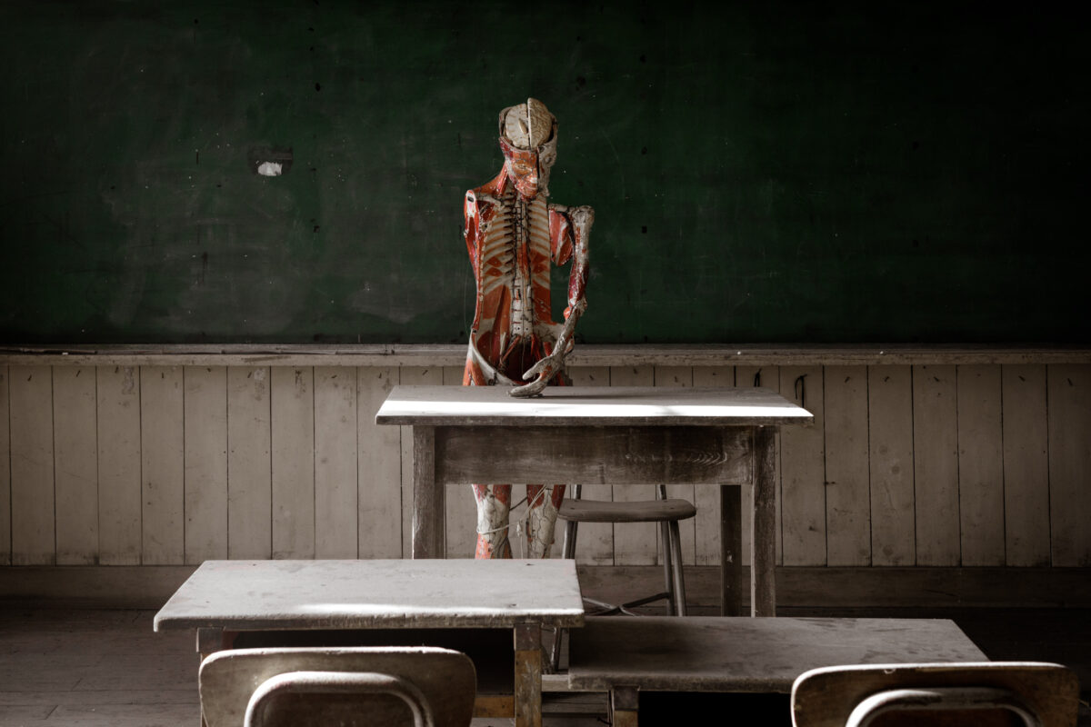 Abandoned classroom with anatomical model behind teacher’s desk and dusty student desks