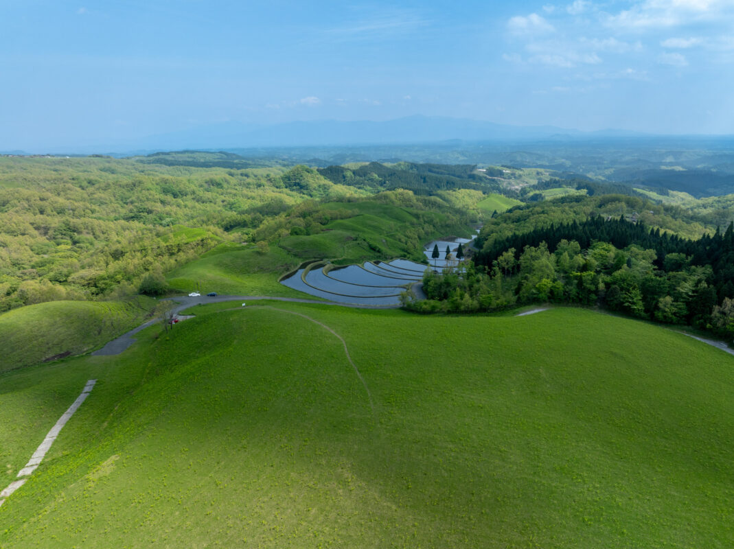 Aerial view of Ogi terraced rice paddies on green hills with forest and distant mountains.